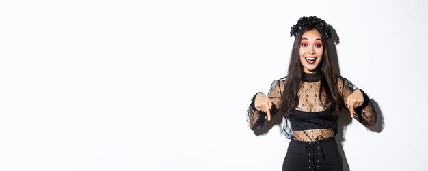 Happy beautiful asian woman in halloween gothic dress pointing fingers down as showing announcement demonstrating your promo standing over white background