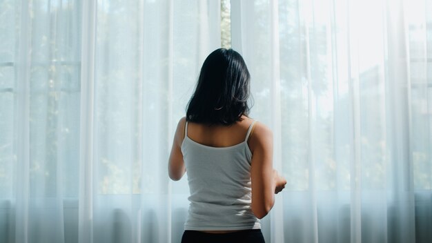 Happy beautiful Asian woman smiling and drinking a cup of coffee or tea near the window in bedroom. Young latin girl open curtains and relax in morning. Lifestyle lady at home .