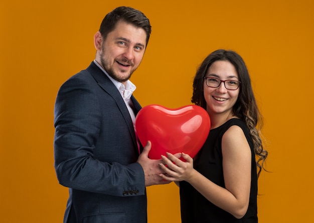 Happy and beautiful couple man and woman with red balloon in heart shape embracing celebrating valentines day over orange wall