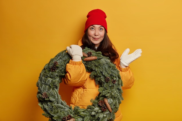 Free photo happy beautiful ethnic woman looks thoughtfully above, wears red hat, warm coat and white mittens, carries green spruce wreath, thinks above celebrating christmas