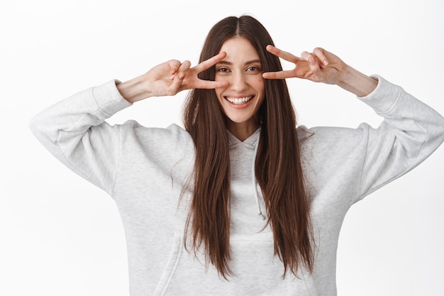 Happy beautiful female model with long healthy straight hair, showing v-sign peace gesture, disco fingers over eyes, smiling broadly, standing against white wall