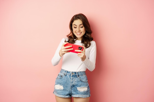 Free Photo happy beautiful girl playing mobile video game, holding smartphone horizontally, watching on cellphone with excited face, pink background