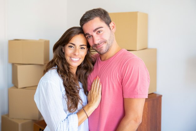 Happy beautiful Hispanic couple standing among carton boxes in their new flat, hugging and looking at camera