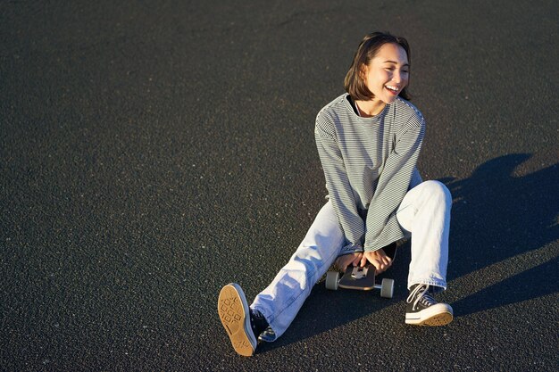 Happy beautiful korean teen girl sits on her skateboard cruising on longboard wearing casual clothes