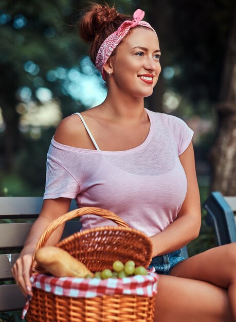 Happy beautiful redhead female wearing casual clothes sitting with a picnic basket on a bench in a park.