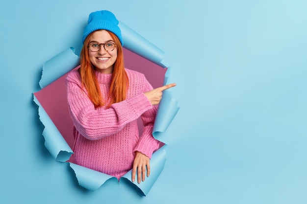 Happy beautiful redhead young woman points right at copy space, wears blue hat and knitted blue jumper