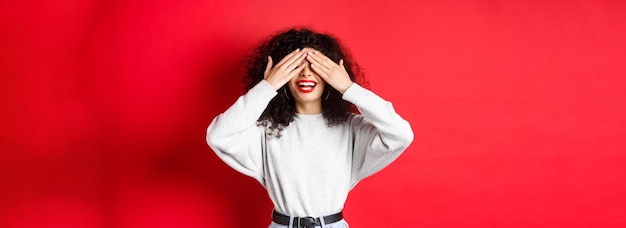 Free photo happy beautiful woman with curly hair and red lips covering eyes with hands and waiting surprise smi