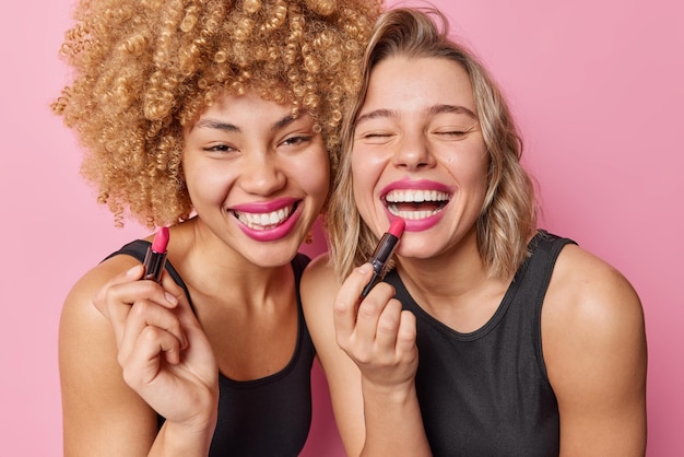 Free photo happy beautiful women apply lipstick undergo beauty procedures stand next to each other dressed in black t shirts isolated over pink background two female friends prepare for party want to look good