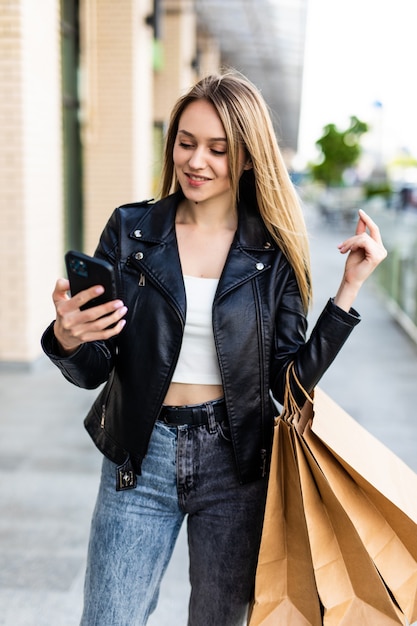 Free Photo happy beautiful young woman with shopping bags, uses a mobile phone, standing in the city