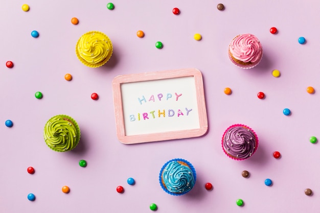 Happy birthday slate surrounded with colorful gems and muffins on pink background