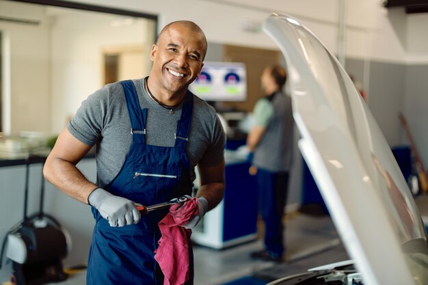 Happy black auto mechanic working at car workshop and looking at camera