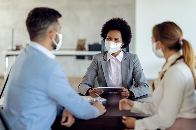 Happy black bank manager wearing face masks while having consultation with a couple in the office