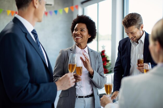 Happy black businesswoman talking to colleagues while drinking Champagne at office party