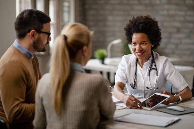 Happy black female doctor talking to a couple and showing them medical test results on a touchpad during consultations at clinic