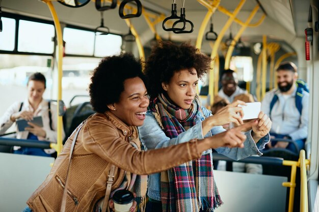 Happy black female friends using smart phone and having fun while traveling by bus