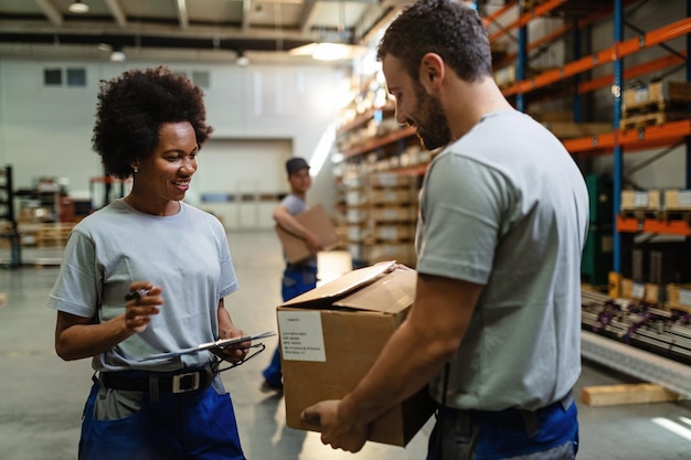 Happy black female worker going through check list and talking to her coworker who is carrying cardboard box in distribution warehouse