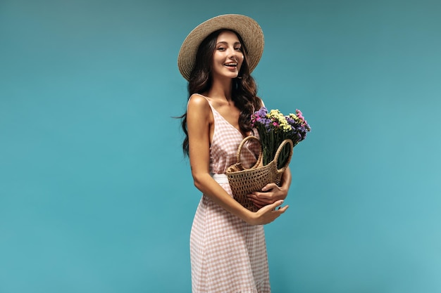 Happy black haired lady in widebrimmed straw hat and long pink and white sundress smiling and holding beautiful bouquet