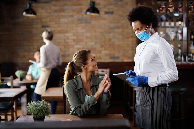 Happy black waitress wearing protective face mask while taking order from customer on a touchpad