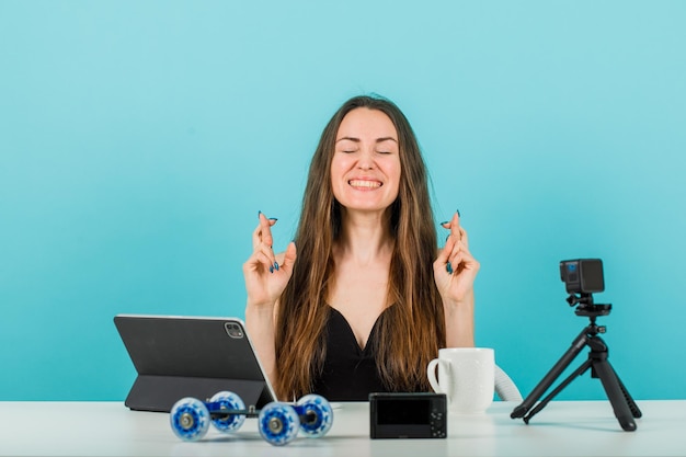 Happy blogger girl is closing eyes by crossing fingers on blue background