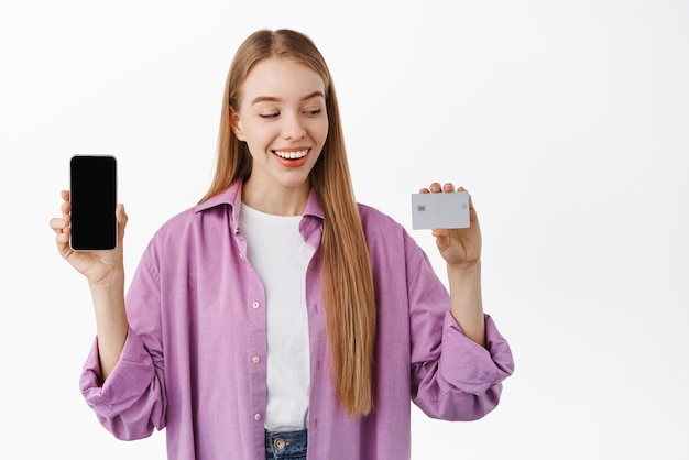 Free photo happy blond girl looking at her bank credit card and showing smartphone app display mobile phone empty screen smiling pleased standing over white background