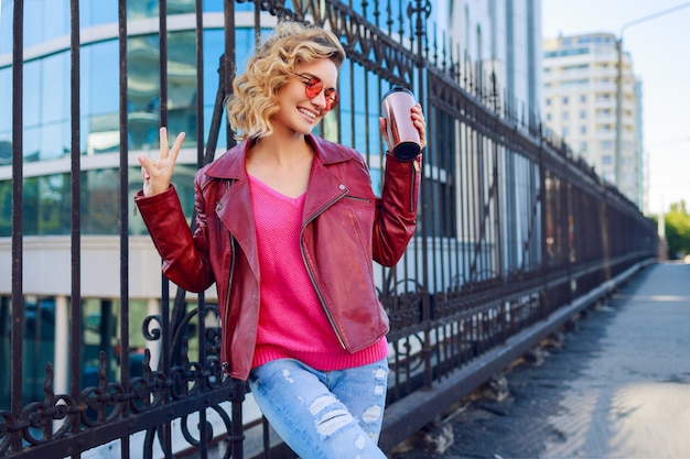 Happy blond woman posing on modern streets, drinking coffee or cappuccino. Stylish autumn outfit, leather jacket and knitted sweater. Pink sunglasses.