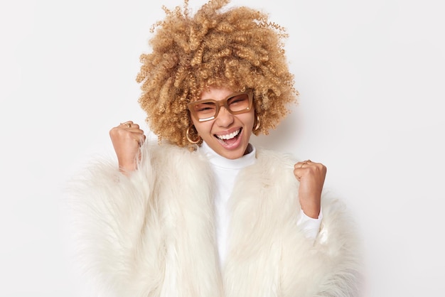 Free Photo happy blonde curly haired young woman shakes fists celebrates success makes yes gesture accepts great offer wears spectacles and white fur coat poses indoor achieves goal yeah we are champions