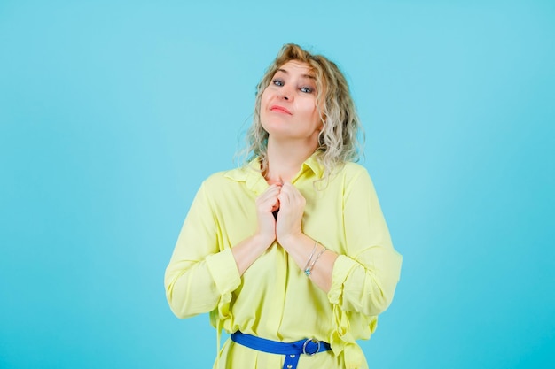 Happy blonde woman is looking at camera by holding fists on chest on blue background