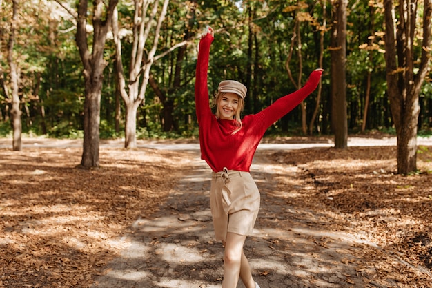 Happy blonde woman in nice red sweater and beige shorts dancing in autumn park. Stylish young woman posing with joy in good weater outdoor.