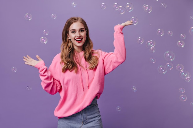 Free photo happy blonde woman in pink sweatshirt playing with bubbles on purple background young curly girl in denim skirt widely smiling on isolated