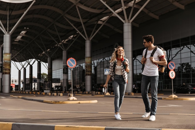 Happy blonde woman talks with her boyfriend Fulllength portrait of cool girl in red headphones and brunette guy in white tee walks with backpacks near airport