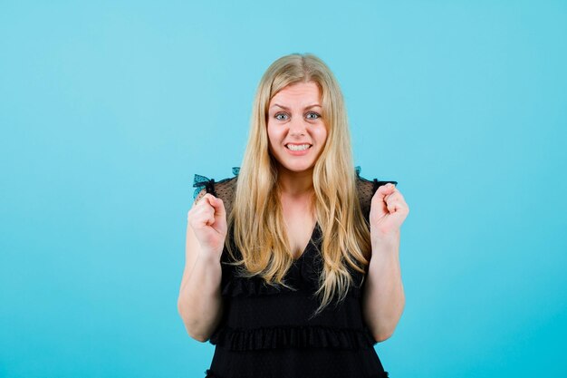 Happy blonde young girl is raising up her fists on blue background