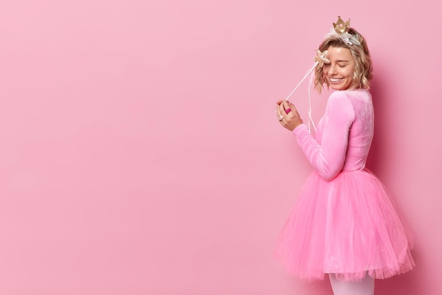 Free photo happy blonde young woman wears festive dress pretends being magical fairy holds wand stands in profile isolated over pink background empty space for your promotional content princess on party