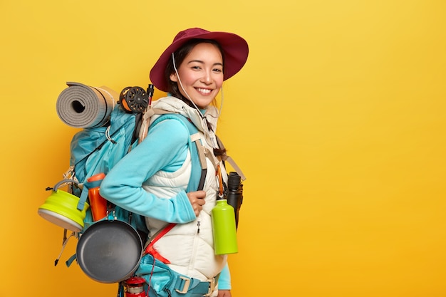 Happy brunette Asian traveler carries big tourist backpack, uses binoculars for journey, stands against yellow wall, wears stylish hat, jumper with vest