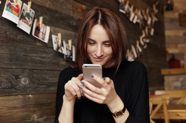 Happy brunette girl in black clothing using free wireless internet connection on smartphone while having rest at restaurant with cozy interior and pictures hanging on wooden wall