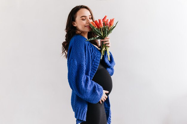 Happy brunette woman in blue cardigan and black dress smells tulips. Charming pregnant lady holds bouquet of flowers on isolated.