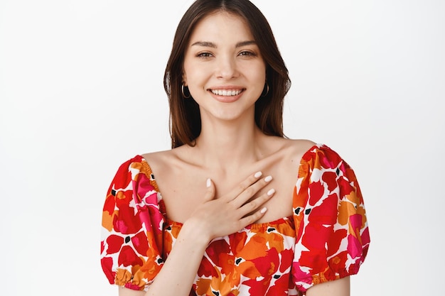 Happy brunette woman holding hand on chest smiling and looking grateful standing in floral dress against white background