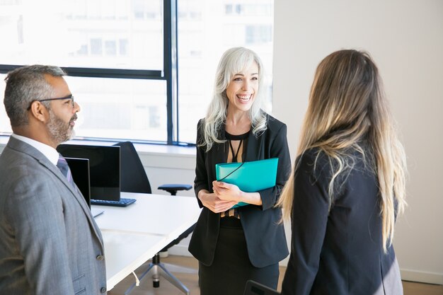 Happy business colleagues wearing suits, standing in office, talking and laughing. Medium shot. Coworkers concept