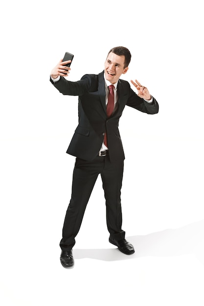 Happy businessman talking on phone over white background in studio shooting. Smiling young man in suit standing and making selfie photo.