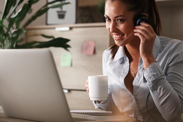 Happy businesswoman drinking coffee while working in customer service center and talking to a customer over the phone