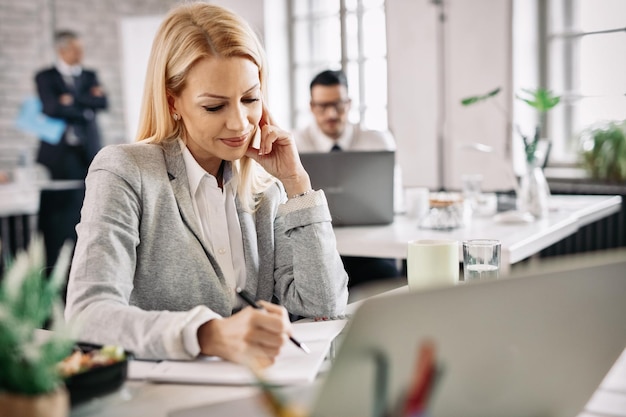 Free photo happy businesswoman working at her desk and taking notes in her note pad there are people in the background