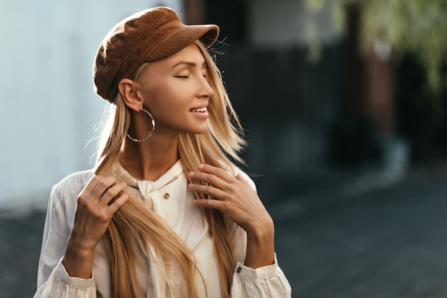 Happy calm young tanned blonde woman in brown cap and white shirt smiles sincerely and poses outside