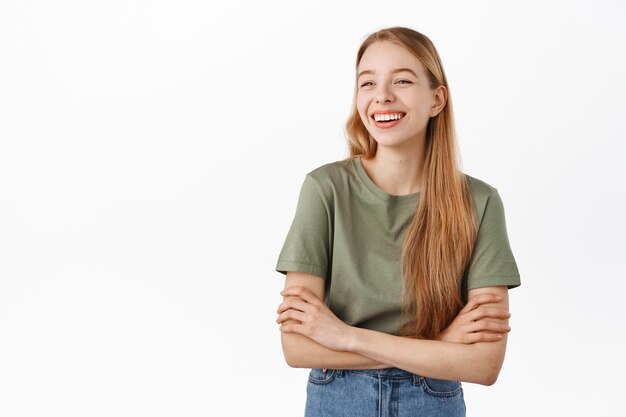 Happy candid girl laughing and smiling, looking aside at advertisement logo with pleased face, standing as if having casual relaxing conversation with friends, white wall