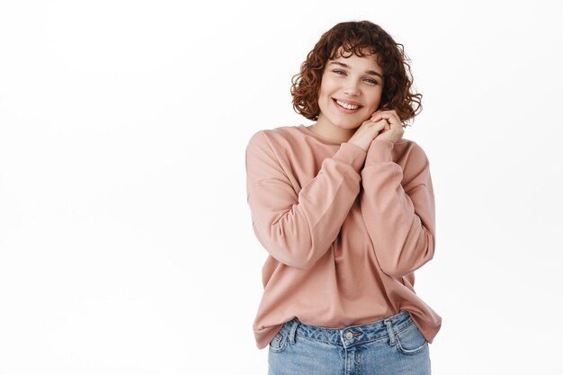 Happy candid girl with beautiful smile, lean head on hands and look at camera with pleased dreamy face, romantic thoughts, standing over white background. Copy space