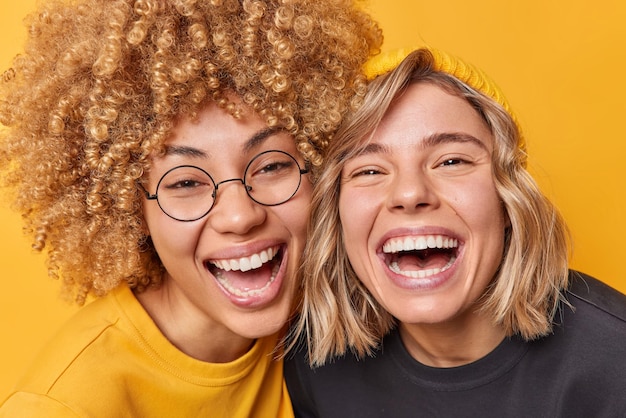 Free photo happy carefree friendly female models laugh joyfully stand next to each other smile toothily have upbeat mood isolated over yellow background people friendship and positive emotions concept