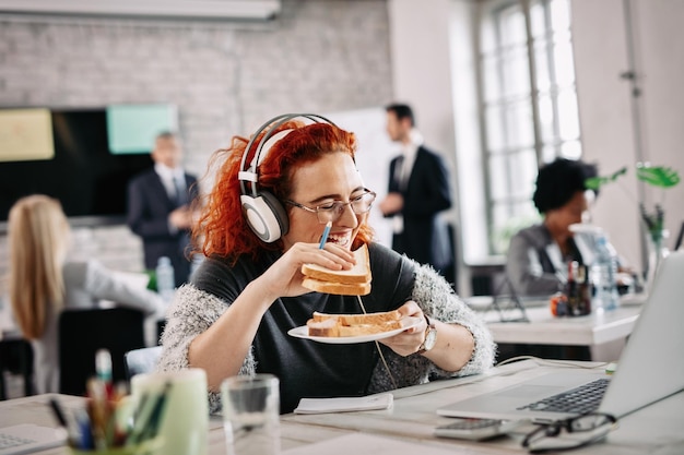 Happy casual businesswoman having fun and laughing while eating sandwich on a break at work There are people in the background