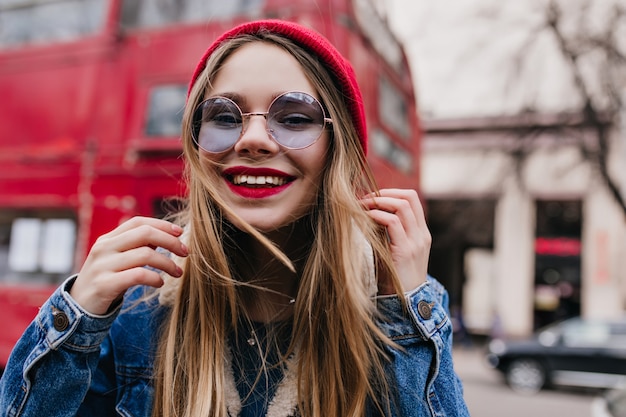 Happy caucasian girl in denim jacket posing on blur street. Outdoor shot of charming white lady in stylish blue glasses.