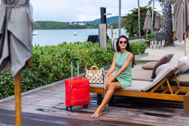 Free photo happy caucasian pretty long hair elegant tourist woman in dress with red suitcase outside hotel