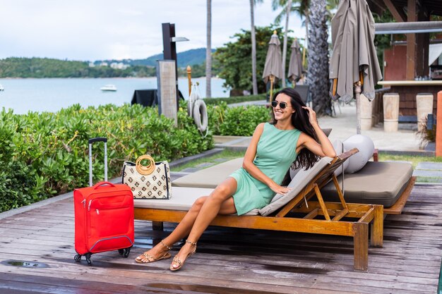 Happy caucasian pretty long hair elegant tourist woman in dress with red suitcase outside hotel