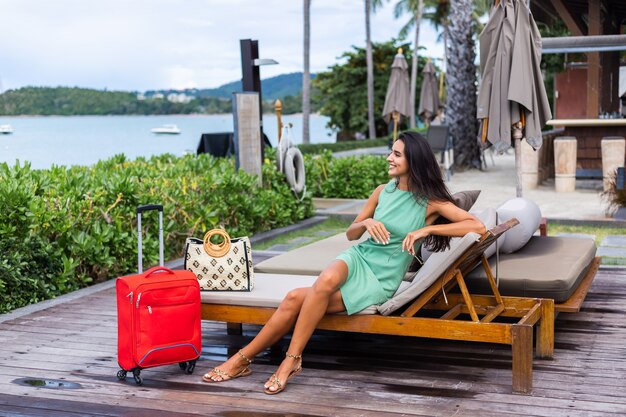 Happy caucasian pretty long hair elegant tourist woman in dress with red suitcase outside hotel