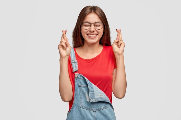 Happy Caucasian woman with dark hair, keeps fingers crossed, prays for good luck, hopes dreams come true has broad smile